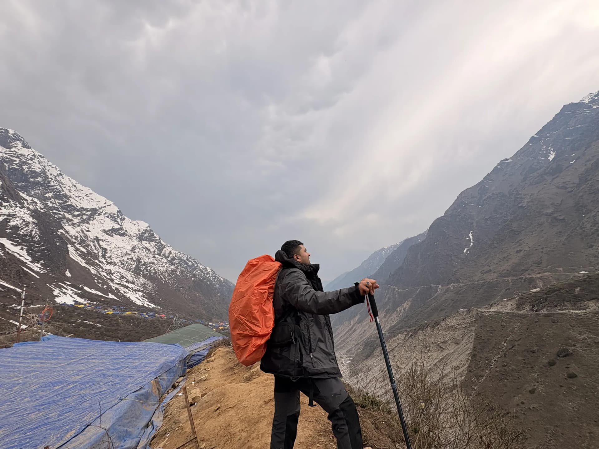 Ganesh Kompella, founder of InnerVeda, on a Himalayan trail with a backpack and trekking pole, gazing towards snow-capped peaks above a glacial valley.